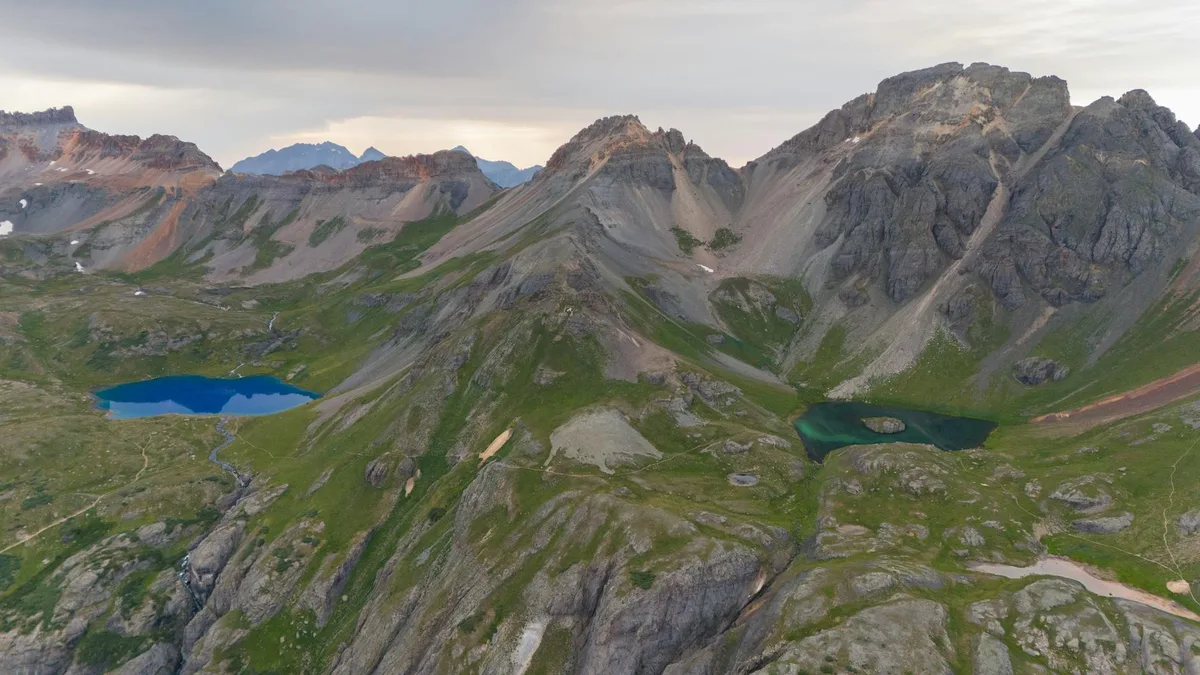 Wide alpine view of three vivid mountain lakes tucked into a rugged basin beneath steep gray peaks, with one bright blue lake on the left and two darker turquoise lakes on the right. Soft morning light and cloudy sky mute the rock faces while green tundra and scattered snow patches spread across the slopes.