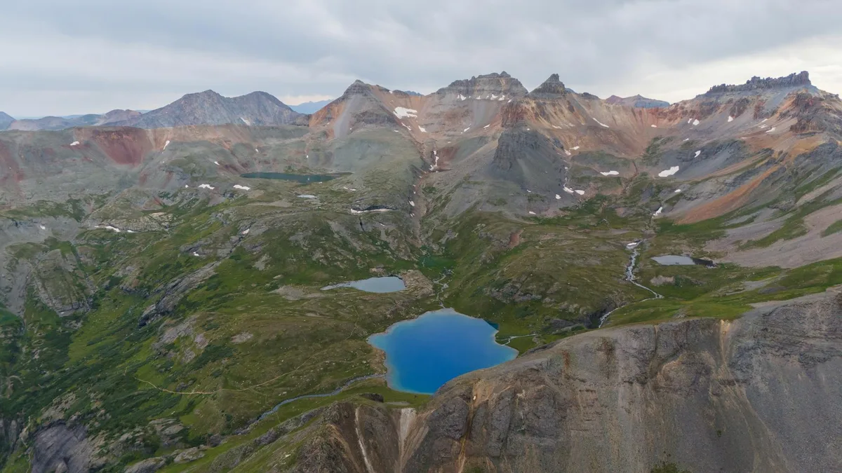 Sweeping aerial view of three turquoise alpine lakes tucked into a rugged basin beneath jagged mountain peaks, with green tundra, gray scree, and small snow patches spread across the high-country slopes. The brightest lake sits in the foreground below a steep rock face under a cloudy morning sky.