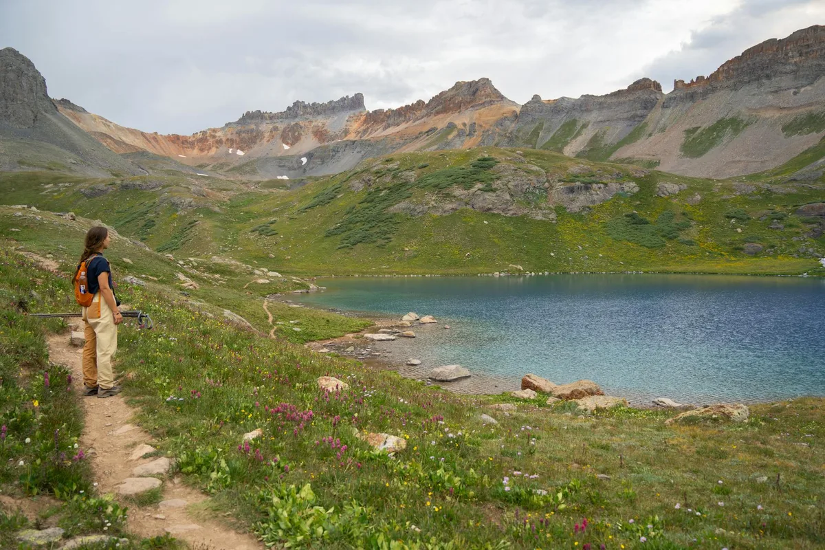 A hiker stands on a narrow dirt trail beside a turquoise alpine lake, with wildflower-speckled meadows in the foreground and the jagged, gray-and-rust slopes of Mount Sneffels rising across the basin. Soft morning light and a pale cloudy sky give the scene a cool, high-altitude feel.