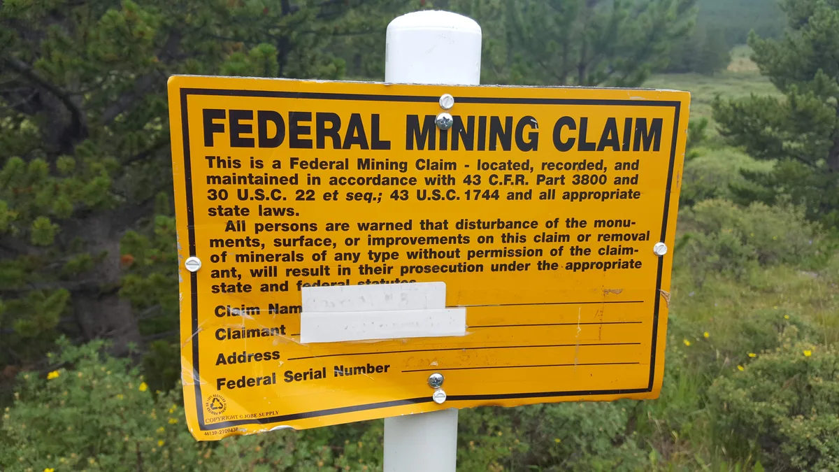 A bright yellow “Federal Mining Claim” sign is bolted to a white post in the foreground, with some blanked-out claim information visible near the bottom. Behind it, a soft-focus green hillside of pines and meadow rises in the mountains.