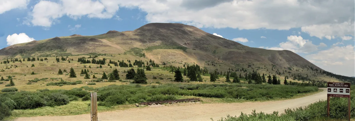A broad alpine mountain rises under a partly cloudy sky, with sparse evergreens scattered across grassy slopes and a dirt road curving through the foreground. A small trail sign and a short stretch of old rail line hint at the route’s historic railroad past.