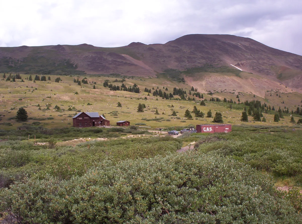 A broad alpine meadow on Boreas Pass stretches toward a dark, rounded mountain under a cloudy sky, with scattered evergreens and low green shrubs in the foreground. A historic wooden building and a red railcar sit near a dirt track below the slope, giving the scene a rustic mining-era feel.