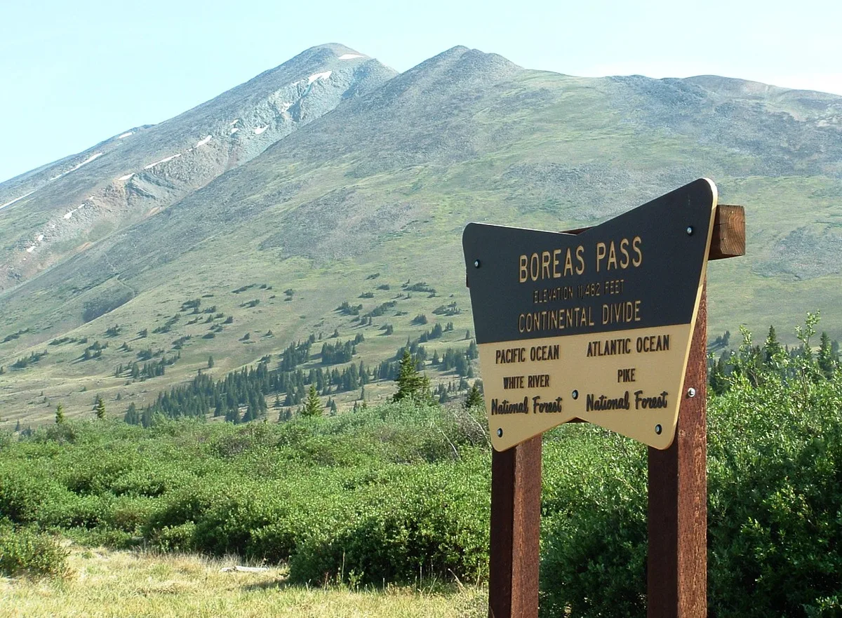 A Boreas Pass sign stands in the foreground, marking the Continental Divide at 11,482 feet, with green alpine slopes and a broad mountain ridge rising behind it. The scene is bright and clear, with scattered evergreens and open tundra under a pale blue sky.