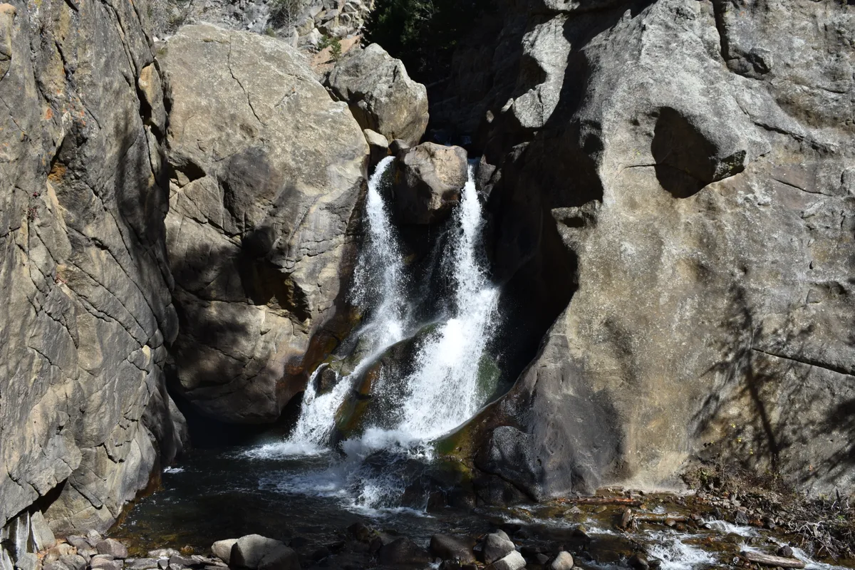 A small waterfall spills in two white ribbons over a dark granite ledge into a shadowed pool, framed tightly by steep canyon walls. Morning light highlights the textured rock face and the foaming water at the base.