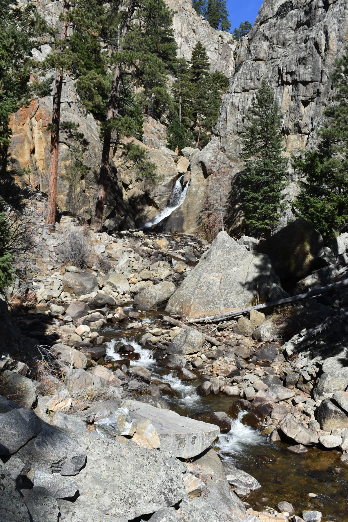 A narrow waterfall drops between pale granite boulders into a rocky creek bed, with white water winding through the canyon below. Tall pine trees and sunlit cliff walls frame the scene under a clear blue sky.