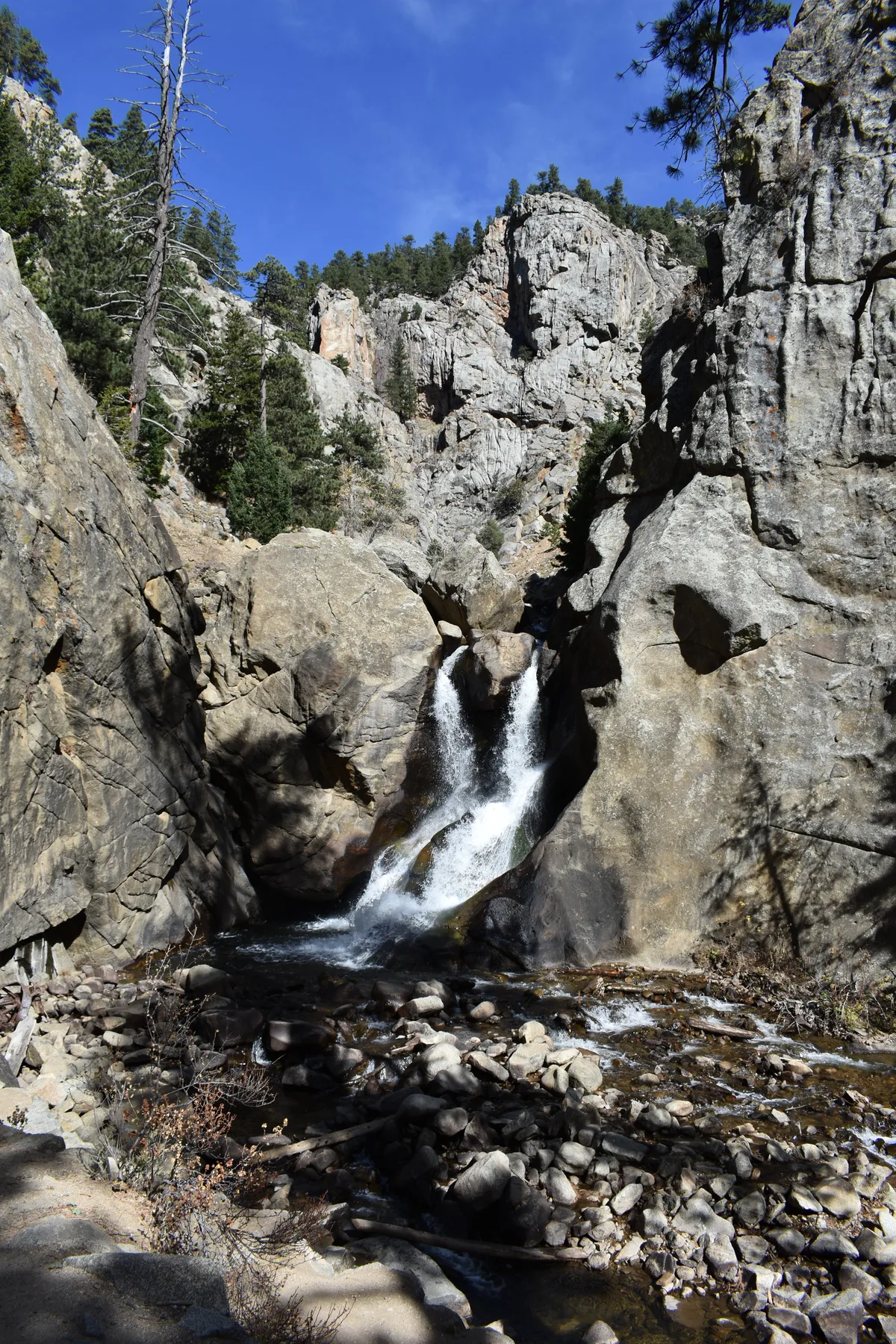 A small waterfall drops between massive gray granite boulders into a rocky pool in a narrow canyon, with pine trees clinging to the cliffs above. Bright morning light and deep shadows emphasize the rough rock textures and the clear stream flowing over stones below.