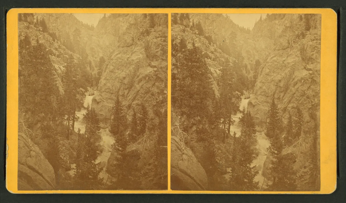 A narrow waterfall drops through a steep granite canyon, framed by dark evergreens and rugged rock walls. The long-exposure view softens the rushing water into bright white ribbons, with warm morning light hazing the distant cliffs.