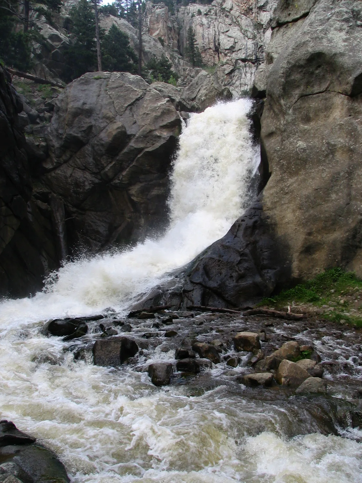 A foaming waterfall surges through a tight granite canyon, tumbling over dark, rounded rocks into a churning creek below. Pine trees cling to the steep cliff walls, with a small patch of green grass at the lower right.