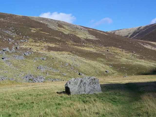A large gray boulder sits in a grassy alpine meadow under a bright blue sky, with rocky brown hills rising behind it. The light is clear and even, emphasizing the open slope and scattered stones across the mountainside.