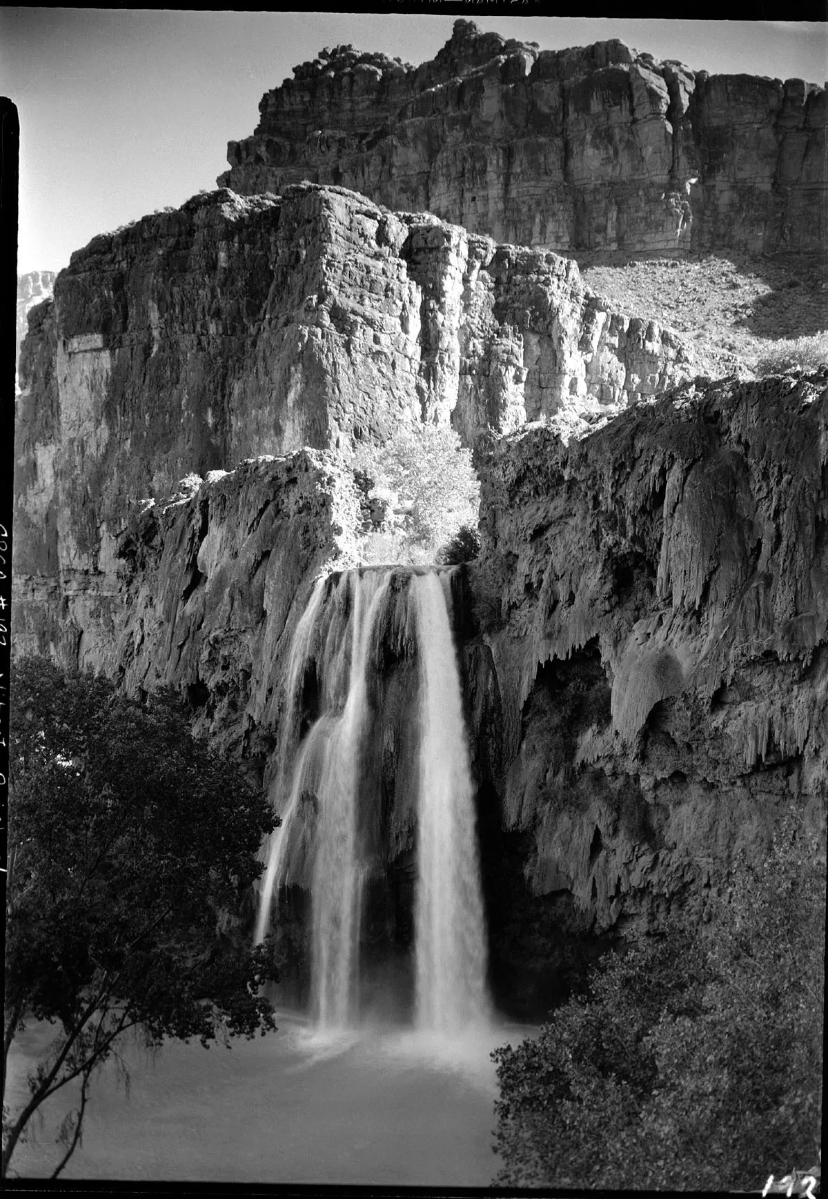 A tall waterfall pours in silky streams over a dark cliff into a misty pool, framed by steep layered canyon walls and sparse trees in the foreground. Bright sunlight catches the rock face, emphasizing the rugged textures and narrow opening above the falls.