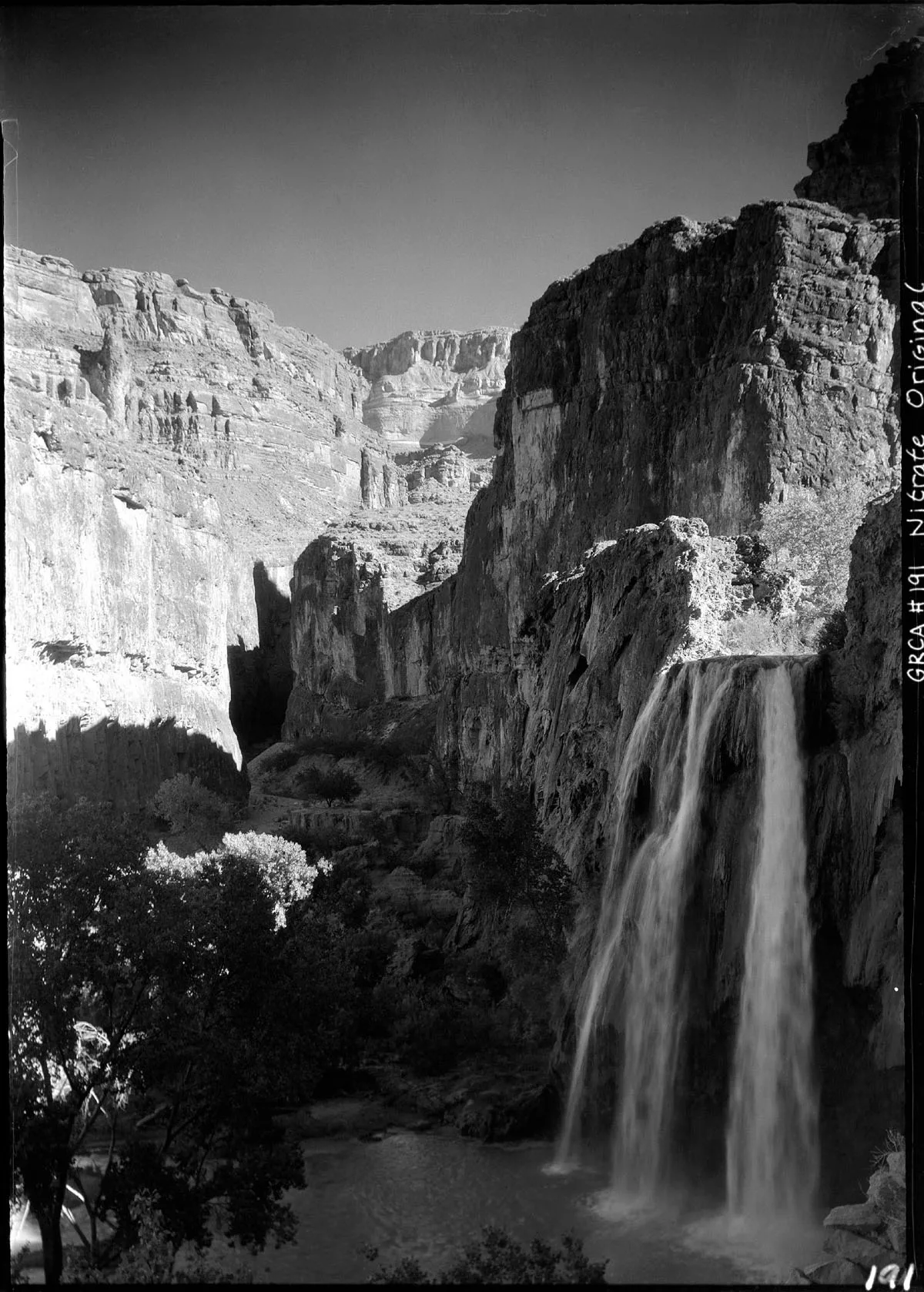 A tall waterfall pours in silky streams off a sheer canyon wall into a shadowed pool, with sunlit layered cliffs rising around a narrow gorge. Dark trees and brush fill the lower foreground, emphasizing the enclosed scale of the canyon.