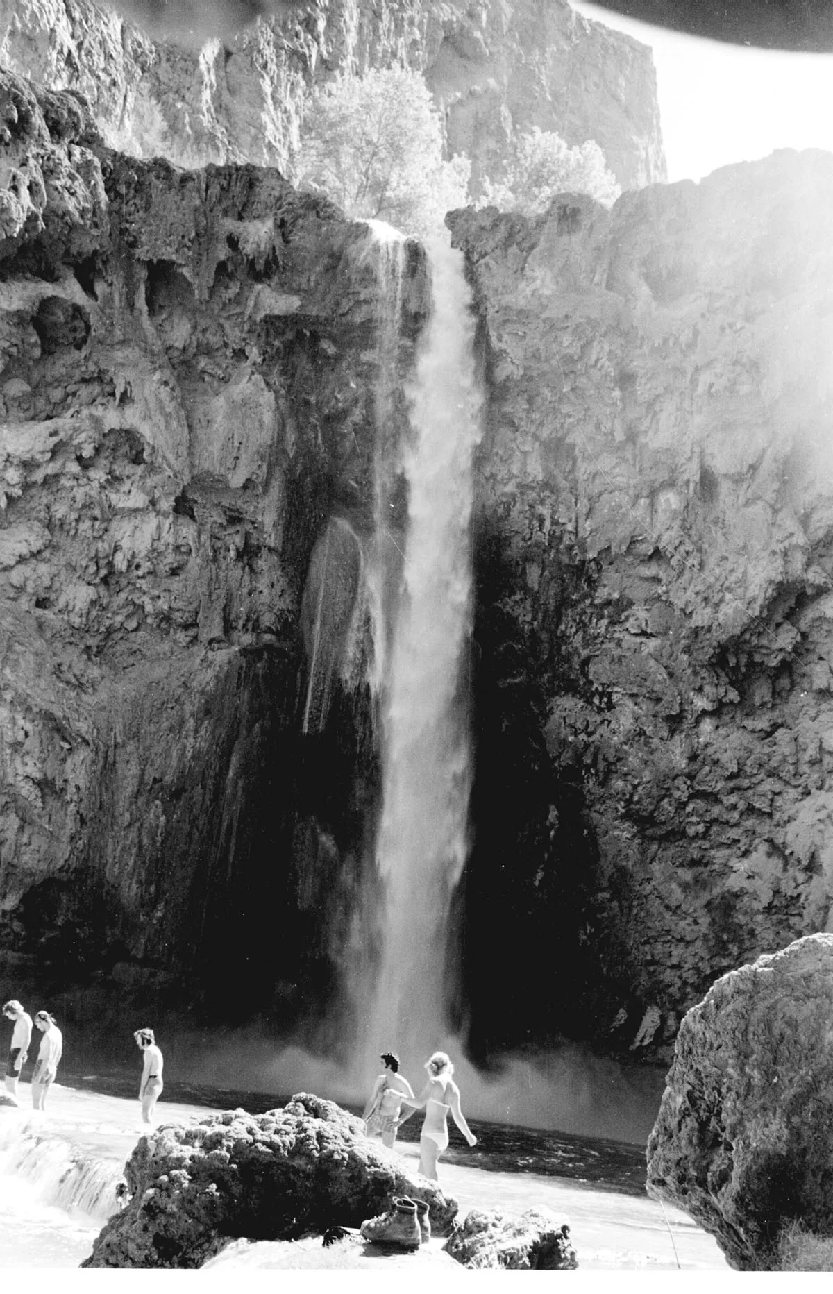 A tall, narrow waterfall plunges into a dark pool between sheer canyon walls, with mist softening the bright upper rim. Several people stand on the wet rocks at the base, emphasizing the scale of the enclosed gorge.