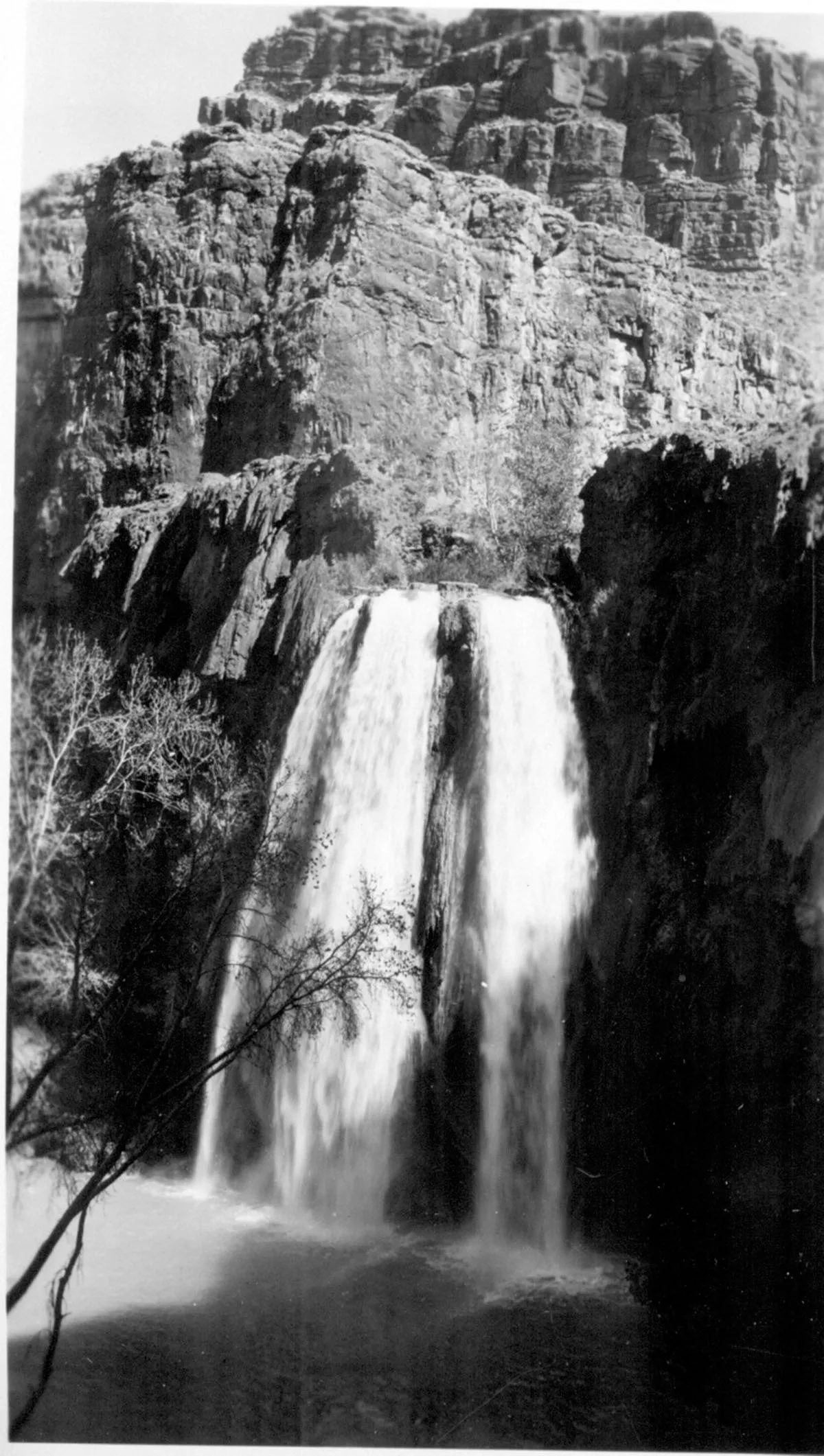 A black-and-white view of a broad waterfall pouring in two silky streams over a dark cliff into a pool below, framed by bare branches and steep layered rock walls. Sunlight catches the pale stone above the falls, while the surrounding canyon stays in deep shadow.
