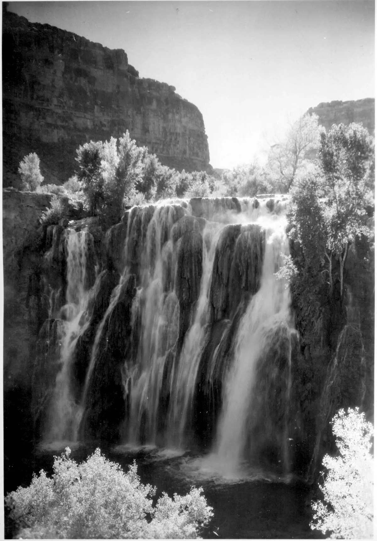 A broad black-and-white waterfall spills in multiple silky streams over a dark rock ledge, framed by bright trees in the foreground and steep canyon walls on both sides. Sunlight washes the upper right of the scene, making the falling water glow against the shaded cliff.