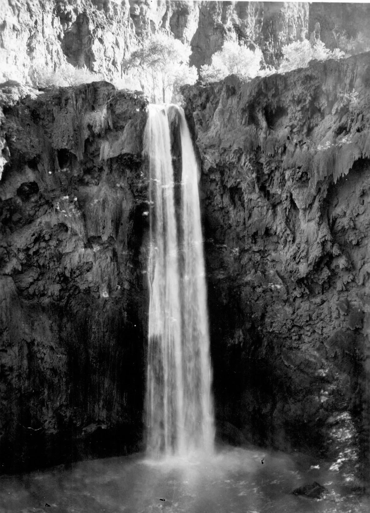 A tall, narrow waterfall drops in a smooth white sheet between dark, sheer canyon walls into a misty pool below. The black-and-white composition emphasizes the enclosed rock face, with bright light catching the rim and sparse trees above the falls.