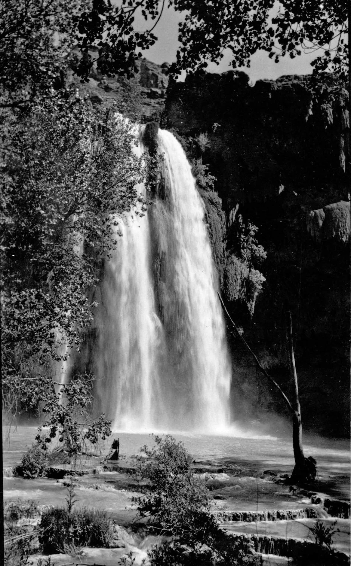 A tall waterfall pours in a smooth white sheet into a shallow river, framed by dense trees and dark canyon walls. Sunlight catches the mist and rippling water, with rocky ledges and low cascades in the foreground.