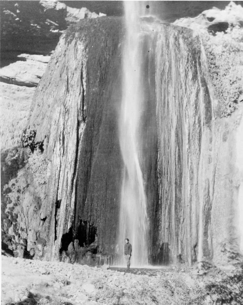 A tall, narrow waterfall pours straight down between sheer canyon walls, turning into a silky white veil in the long exposure. A person stands at the base for scale, emphasizing how massive the dark, striated rock face is around the falls.