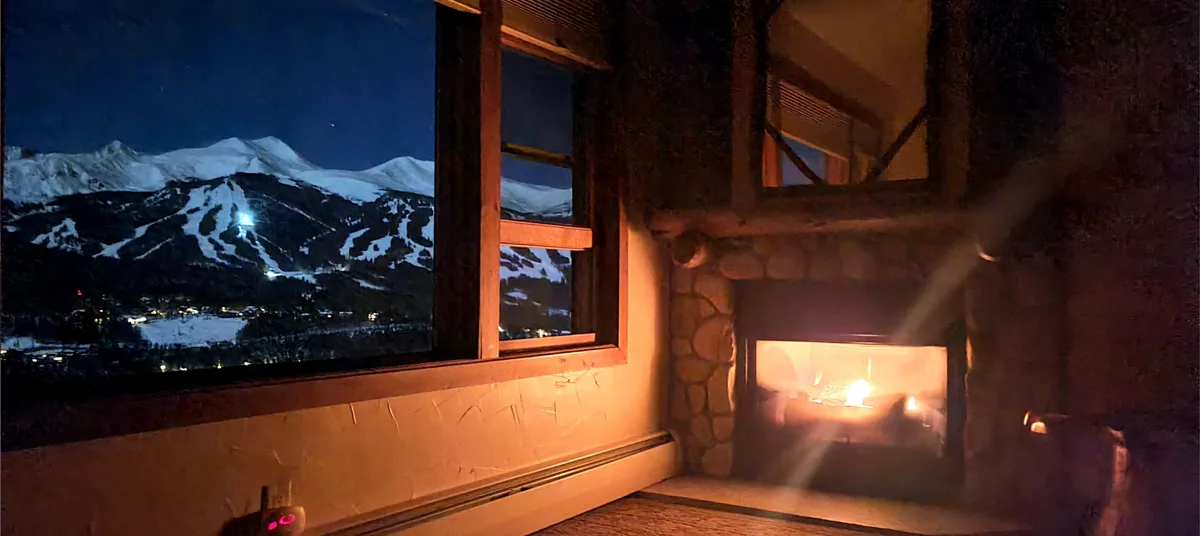 A cozy lodge room glows with a fireplace on the right and a large window on the left framing snow-covered Breckenridge ski slopes under deep blue twilight. Warm firelight and the dark wooden interior contrast with the icy mountain view outside.