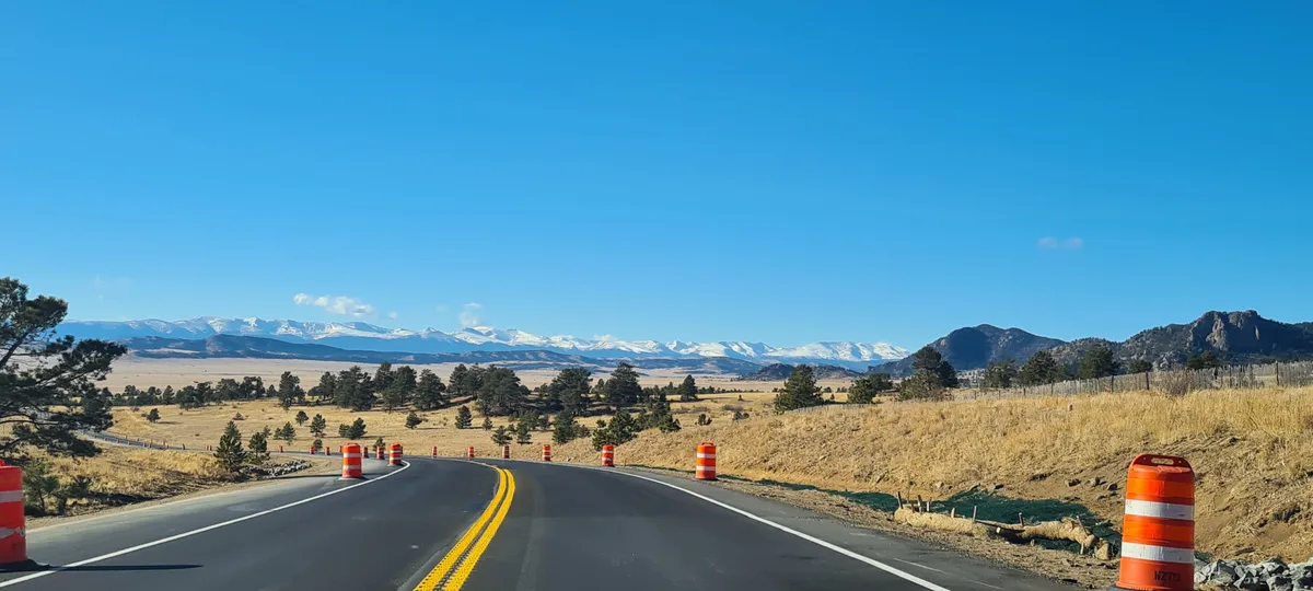 A curving two-lane road leads through dry golden grassland and scattered pines, with orange construction barrels lining the shoulder under a clear blue sky. Snowcapped mountains stretch across the horizon, creating a wide open high-country landscape.