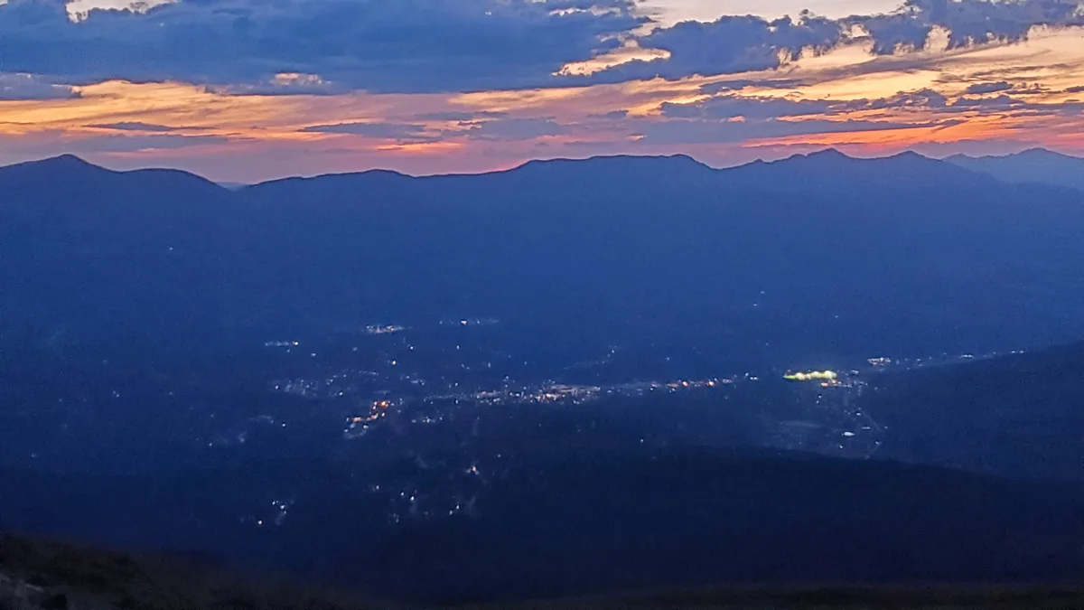 Breckenridge sits in a dark blue mountain valley at dusk, with scattered town lights glowing below a silhouette of the Tenmile Range. Streaks of pink and gold sunset light the clouds above the ridge, while the foreground falls into deep shadow.