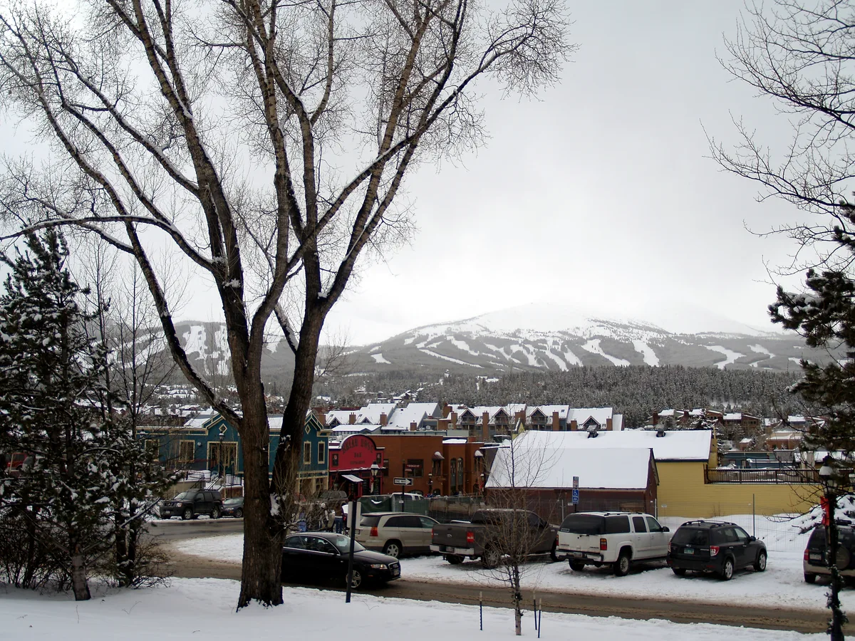 Snow-covered Breckenridge Main Street is seen from an elevated vantage point, with colorful Victorian storefronts and parked cars framed by bare trees in the foreground. Behind the town, the Tenmile Range rises under an overcast winter sky, with ski runs faintly visible on the mountain.
