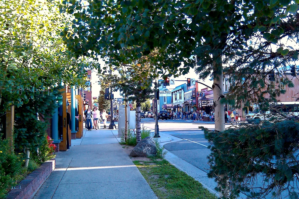 Tree-shaded sidewalk along Breckenridge’s main street, with colorful Victorian-style storefronts, pedestrians, and a busy intersection in bright daylight. A line of leafy branches frames the view, softening the streetscape and drawing attention to the historic downtown buildings.