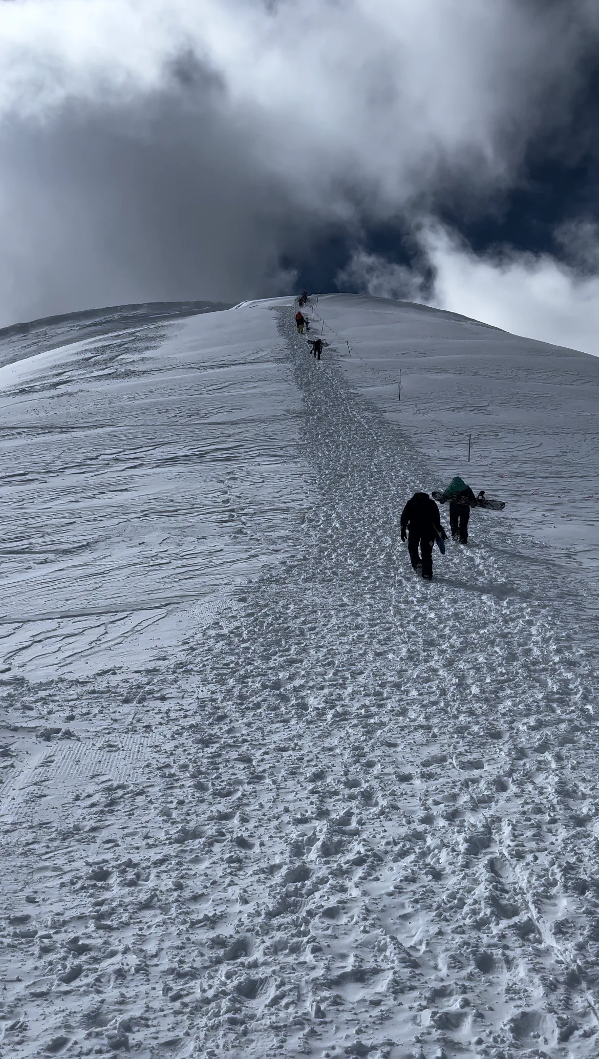 Hikers climb a narrow, trampled snowy ridge toward a rounded summit, with wind-sculpted snow and exposed footprints leading the eye upward. Dark, fast-moving clouds fill the sky above the stark white slope, creating a dramatic high-alpine scene.