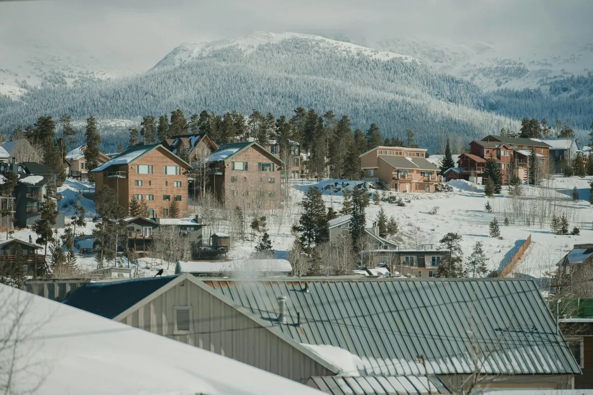 Snow-dusted mountain homes with peaked roofs fill the middle ground, backed by a steep, pine-covered ridge and a pale winter sky. The foreground is cut by the greenish metal roof of a building, creating layered bands of rooftops, trees, and snow.