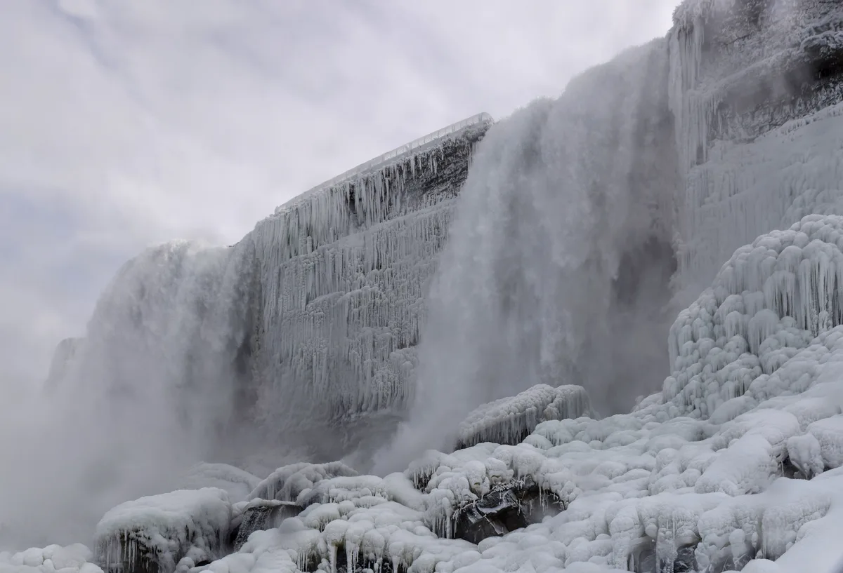 Bridal Veil Falls plunges through a gray winter haze, with thick mist billowing across the frame and the rock face coated in heavy ice and snow. The historic power plant sits along the rim above the frozen curtains of water, while rounded ice formations fill the foreground.