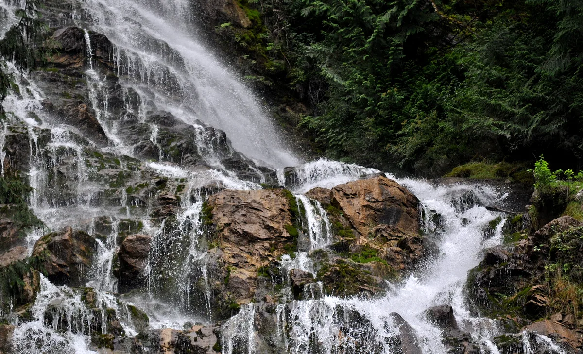 White water spills in multiple thin ribbons over dark, jagged rock ledges, creating a misty cascade with long-exposure blur. Dense green forest frames the falls, and wet tan boulders anchor the lower foreground.