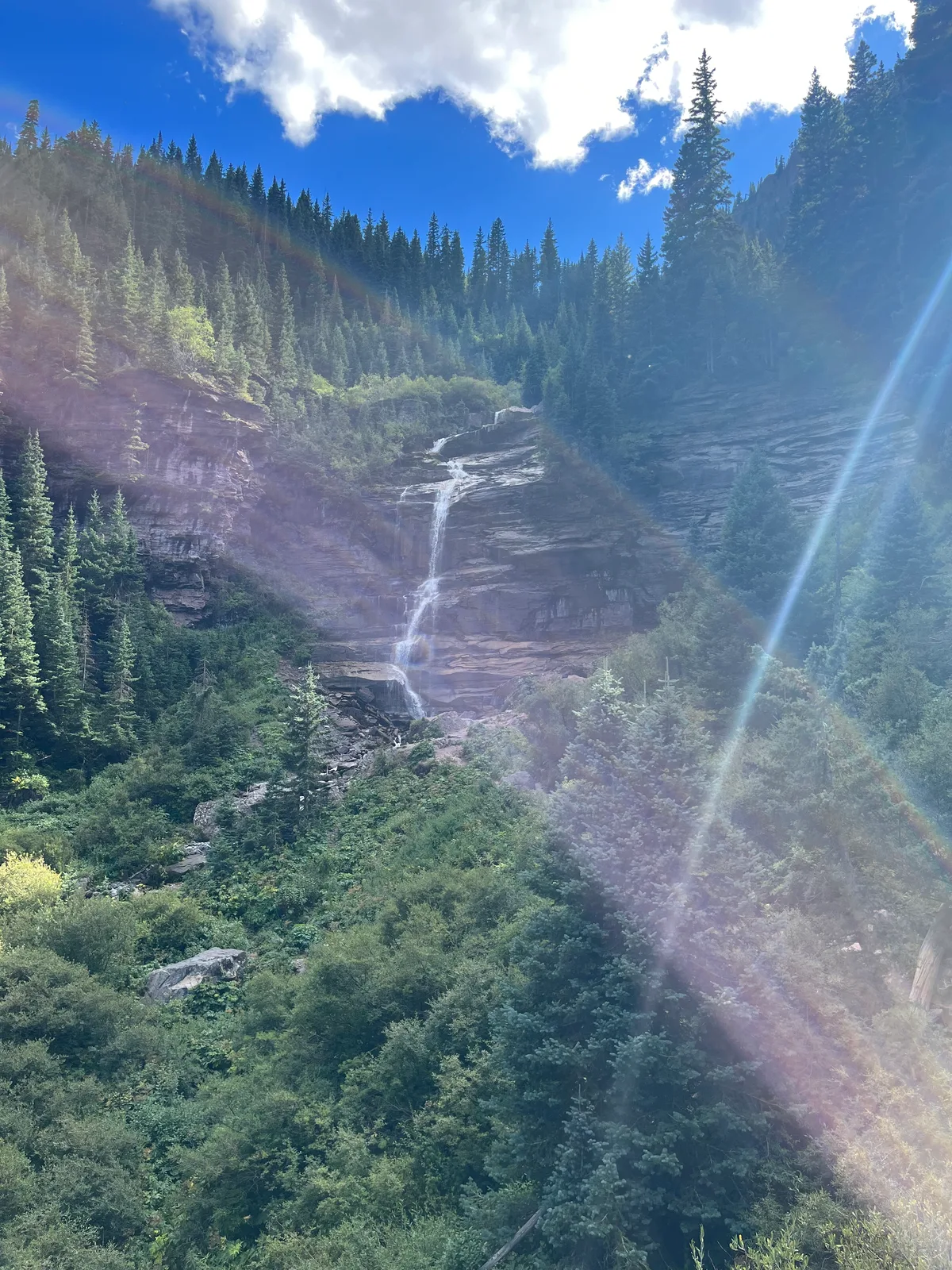 Tall Bridal Veil Falls drops in a thin white ribbon down a dark rock cliff, surrounded by dense evergreen slopes in Telluride’s box canyon. Bright morning light and lens flare streak across the scene beneath a vivid blue sky with scattered clouds.