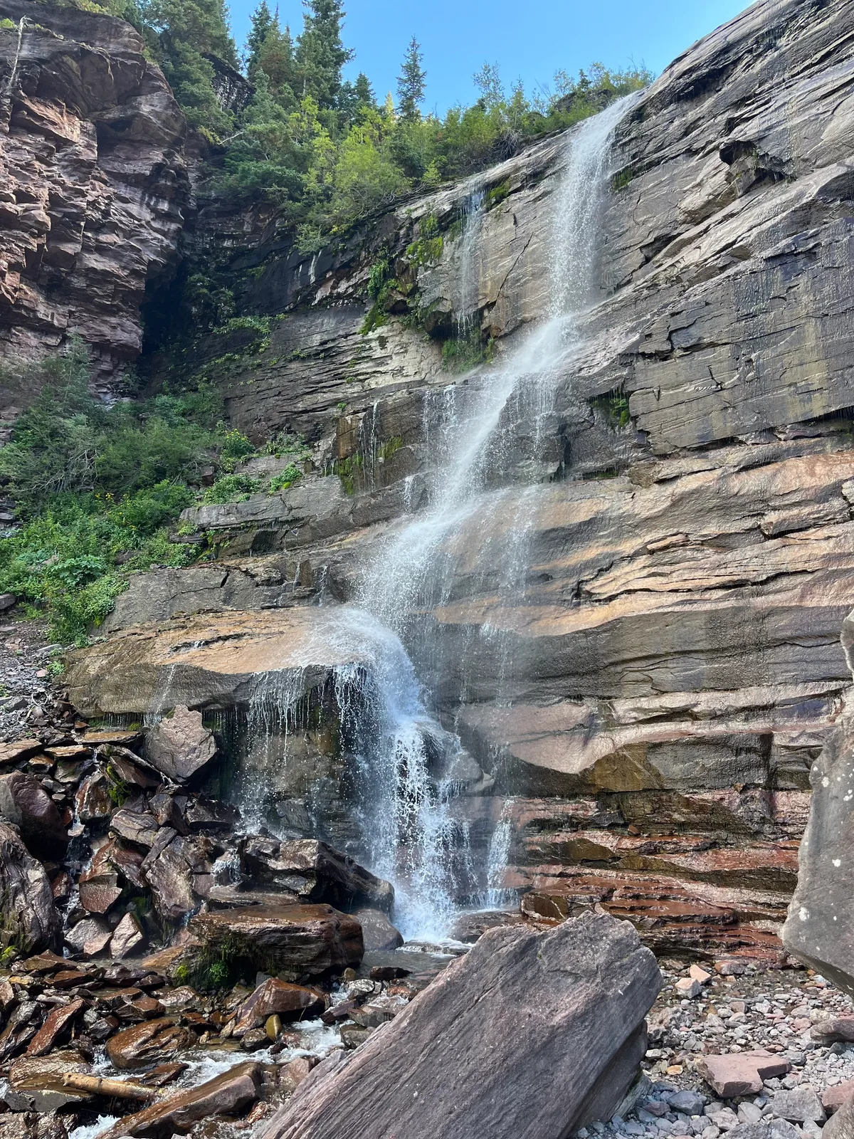 A slender waterfall spills down a steep gray rock face in bright morning light, breaking into mist as it drops into a boulder-strewn basin. Evergreen trees and a clear blue sky frame the cliff, with a large weathered log in the foreground.