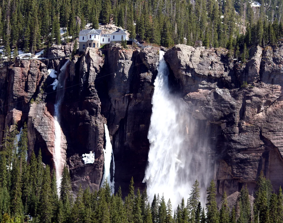 Bridal Veil Falls drops in a tall white plume over a sheer cliff, with mist drifting down beside a smaller ribbon of water. At the top, the historic power plant sits on the rocky rim above dense evergreen forest and lingering patches of snow.