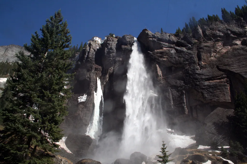 Bridal Veil Falls plunges in a bright white column over dark, rugged cliffs, with mist rising at the base and patches of lingering snow around the rocks. A small historic power plant sits on the rim above the falls, framed by evergreen trees under a clear blue sky.