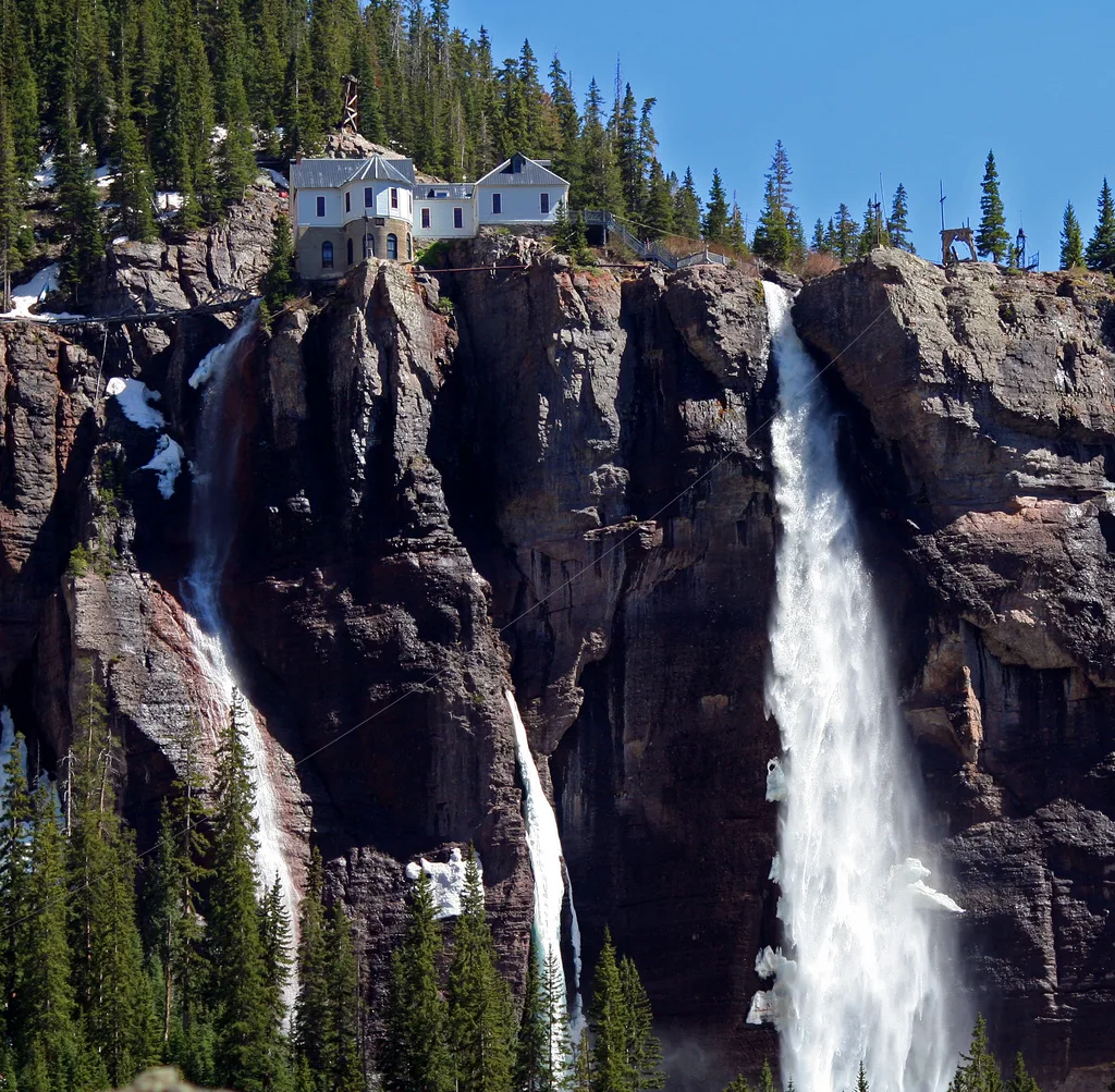 Bridal Veil Falls plunges in multiple white ribbons over a dark rock cliff, with the historic power plant perched at the top amid evergreen trees. Bright morning light and patches of lingering snow emphasize the steep canyon and misty spray at the base of the falls.