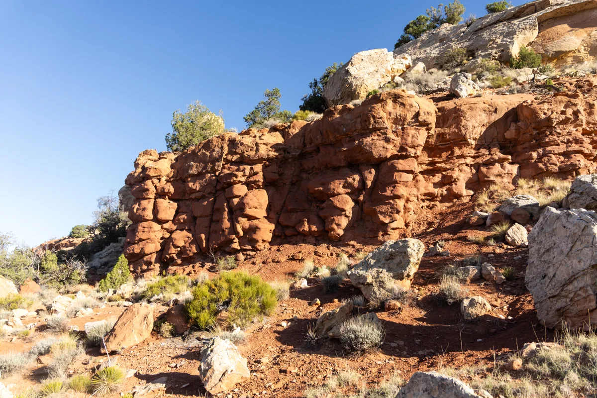 Golden sunlight washes over a red sandstone canyon wall strewn with boulders and scrubby sagebrush, with pale rock ledges capping the bluff against a clear blue sky. The warm light brings out the rough, layered texture of the cliff and the dry, sparse desert landscape.