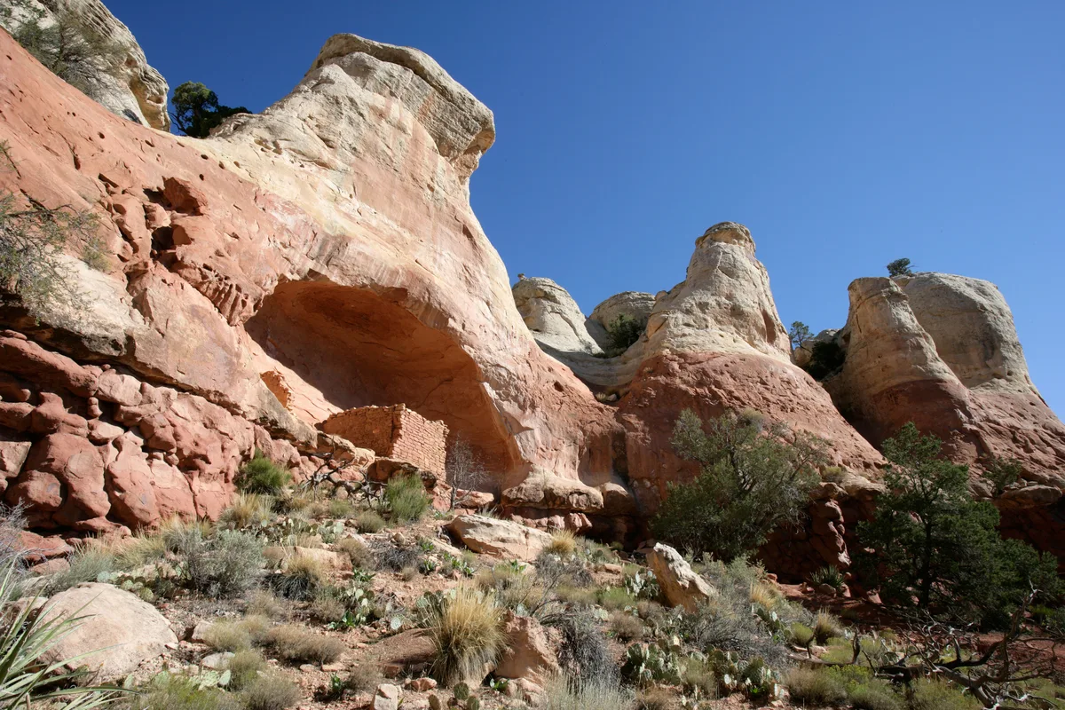 A small ancient stone ruin sits tucked beneath a broad sandstone alcove, surrounded by red-and-cream cliff walls and sparse desert scrub. The low-angle composition emphasizes the towering rock formations against a clear blue sky, with warm light bringing out the layered textures of the canyon.