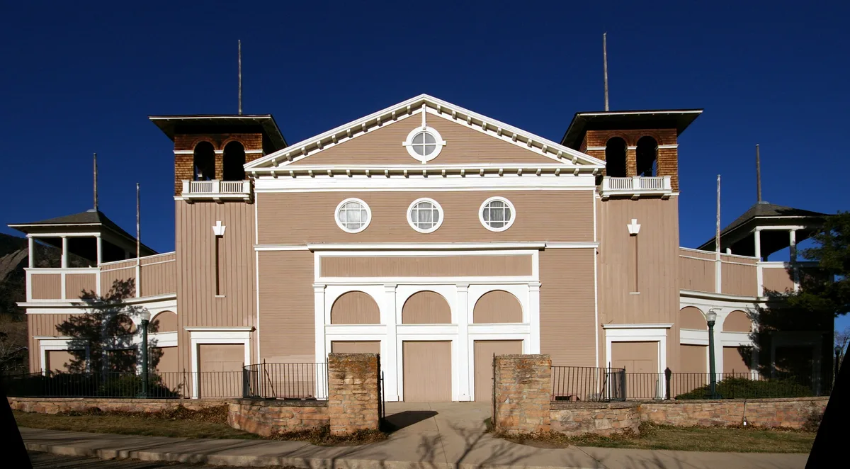 Historic Colorado Chautauqua Auditorium fills the frame, centered against a deep blue sky, with tan wood siding, white trim, round windows, and twin towered wings. The front entrance sits behind a low stone wall and iron fence, with long shadows suggesting late-day light.