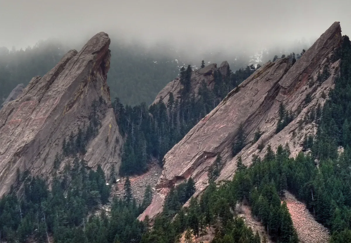 The Flatirons rise in steep, jagged sandstone slabs above a dense evergreen forest, with low clouds and mist softening the ridgeline in the background. The scene feels cool and moody, with muted light emphasizing the rock texture and dramatic angle of the peaks.