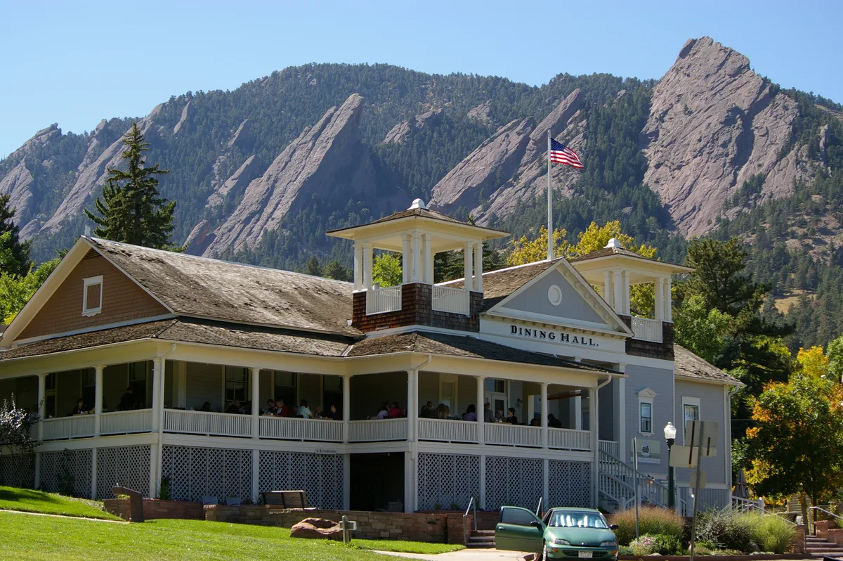 Historic Chautauqua Dining Hall sits in the foreground with wraparound porches, white trim, and an American flag on top, backed by the steep, sunlit Flatirons. A green lawn and a parked car lead into the scene, with late-day light highlighting the gray-blue rock and surrounding trees.