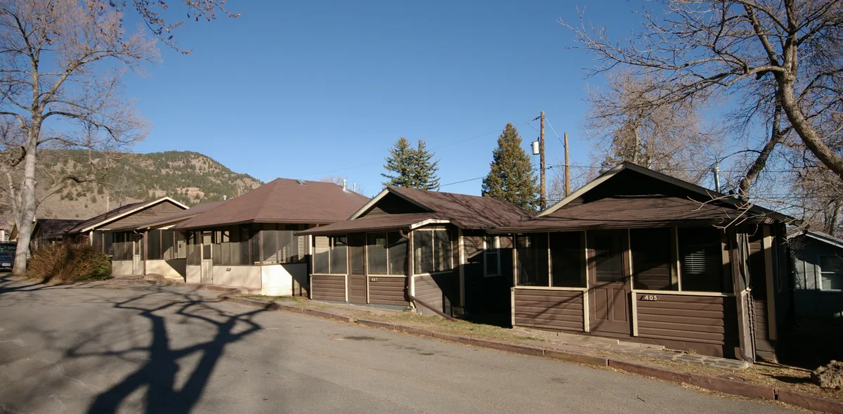 Historic brown cabins with screened porches line a quiet paved drive at Chautauqua Park, with leafless trees framing the scene and the Flatirons rising in the background. Clear blue sky and low golden light cast long shadows across the road and cottages.