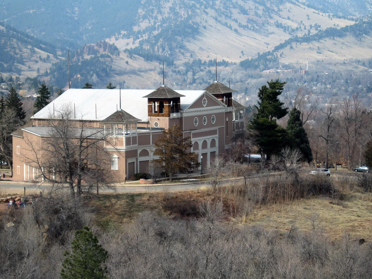 Historic Chautauqua Auditorium sits among leafless trees and grassy slopes, its pale roof and twin towers standing out against the muted foothills. The Flatirons rise softly in the background under hazy daylight, giving the scene a quiet early-spring feel.