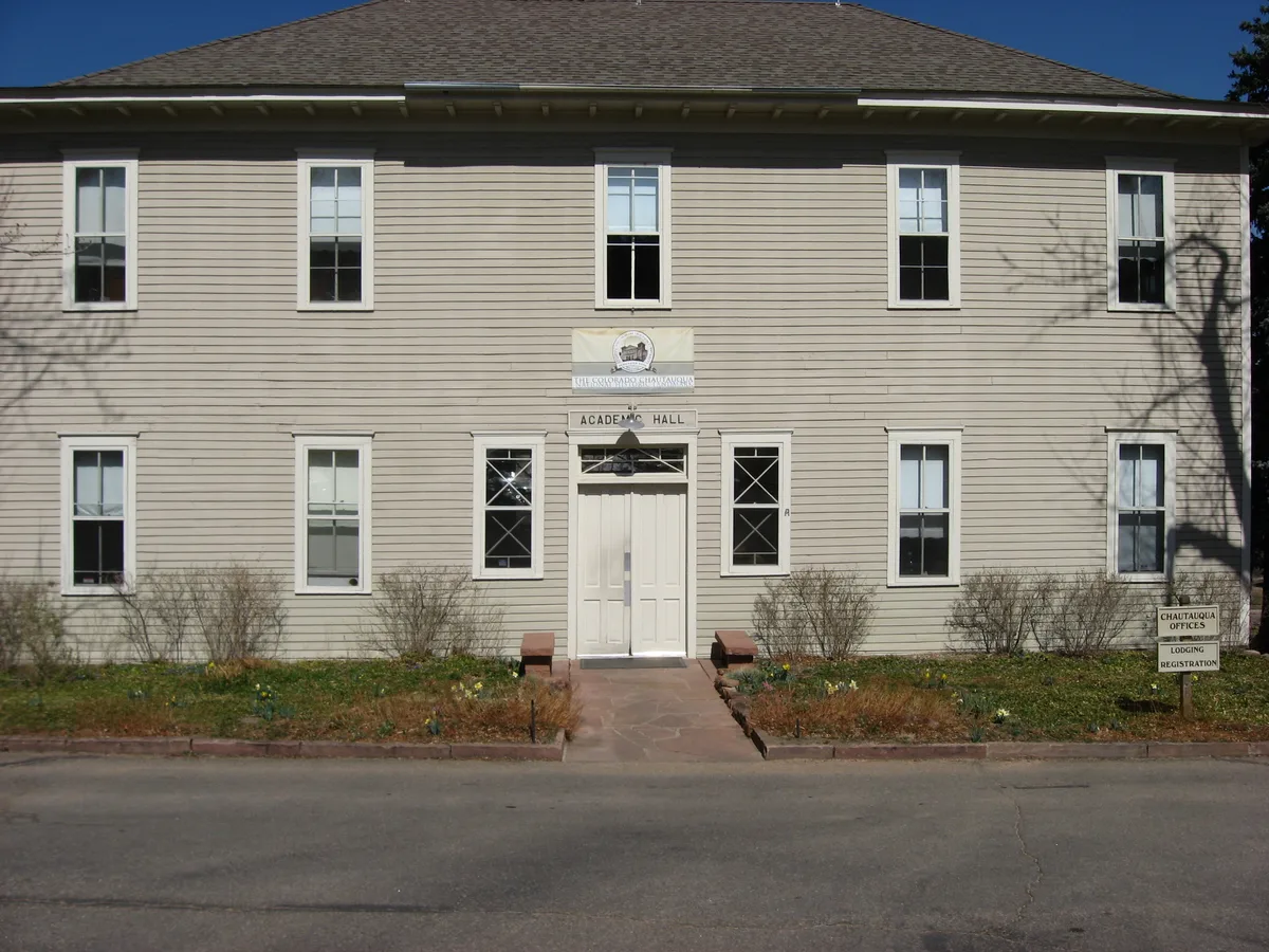 Historic Academic Hall at Colorado Chautauqua fills the frame, with a beige clapboard facade, symmetrical windows, and a centered white double door beneath the hall sign. Bare shrubs and a narrow path front the building, with soft tree shadows falling across the wall in bright daylight.