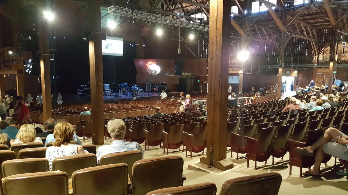 Inside the Chautauqua Auditorium, rows of brown seats fan out beneath exposed wooden beams and tall support posts toward a dimly lit stage with instruments set up for a performance. Scattered attendees sit and chat in the warm indoor light, giving the historic hall a relaxed, anticipatory feel.