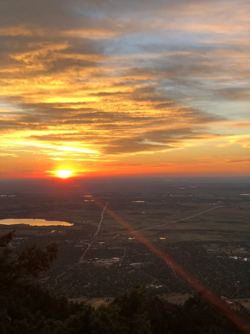 Sunrise glows orange and gold over the distant plains, with layered clouds catching the early light above a hazy horizon. Dark pine silhouettes frame the foreground, and a faint red lens flare cuts diagonally across the view.