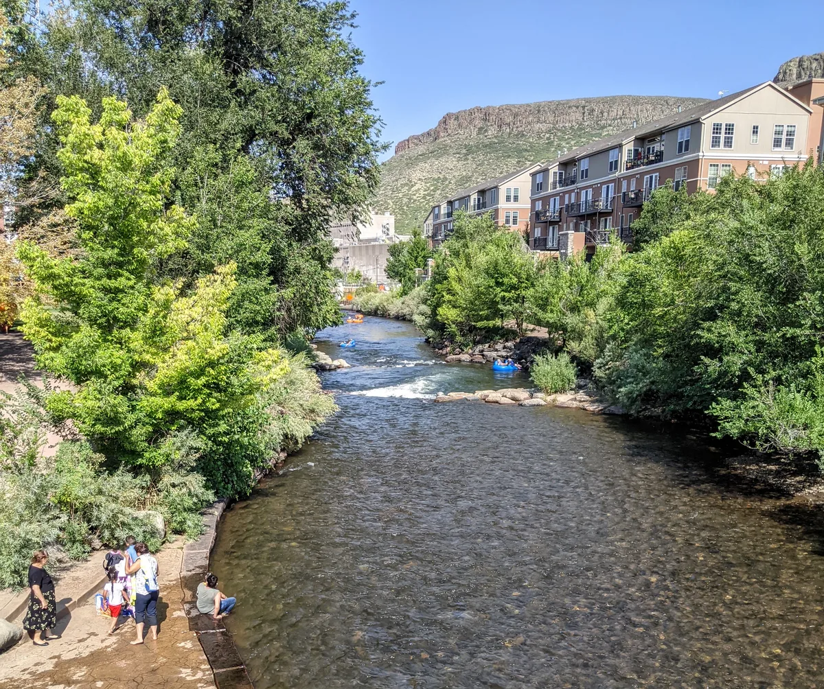 Clear Creek flows between leafy banks and apartment buildings, with tubers drifting through the water and people standing along the paved edge in the foreground. South Table Mountain rises behind the canyon under a bright blue morning sky.