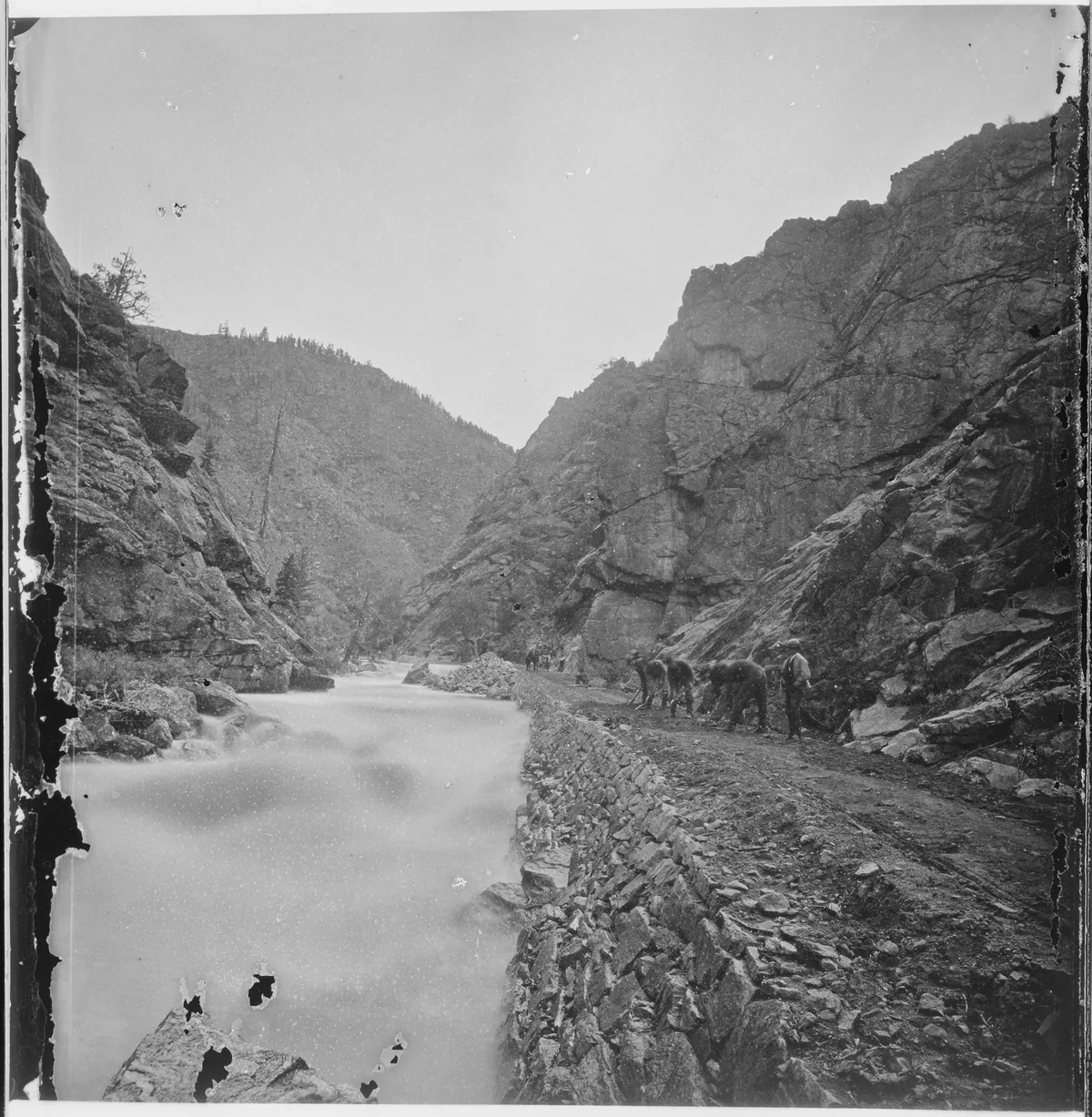 Long-exposure water fills the narrow canyon channel, turning Clear Creek into a smooth, pale ribbon between steep, jagged rock walls. A dirt path and low stone embankment run along the right side, where a small group of people stands beside the rushing creek under a bright, hazy sky.