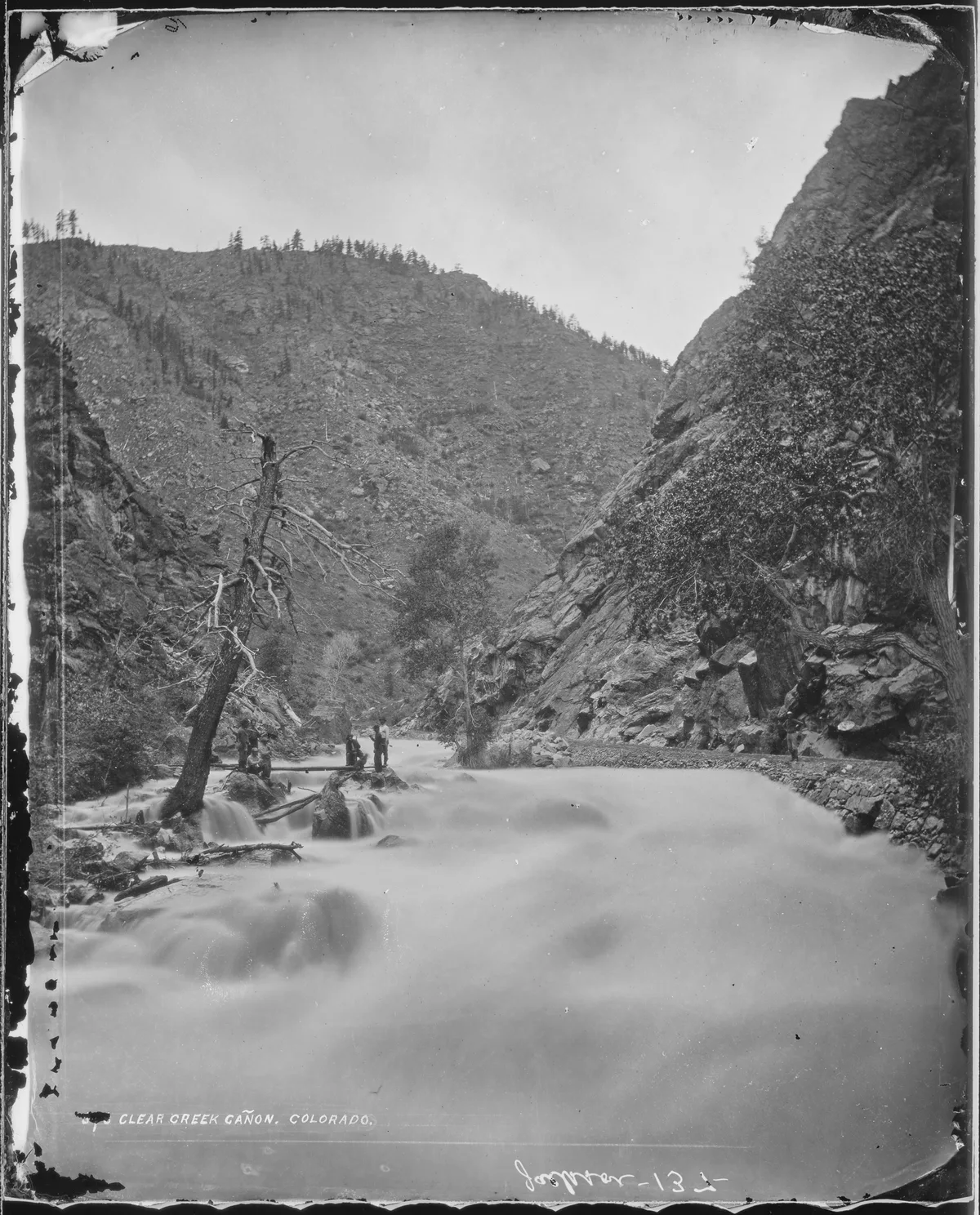 Long-exposure water blurs through Clear Creek Canyon between steep rock walls, with a few people standing on boulders near the creek and a bare tree leaning over the stream. The canyon narrows into a rugged V-shaped gap under a bright sky, with sparse trees lining the rocky slopes.