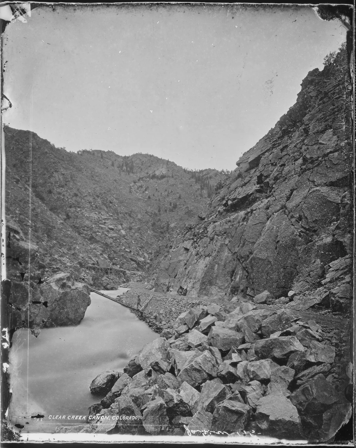 Long-exposure water flows past a large boulder in a narrow canyon, with steep rocky walls rising on both sides and scattered boulders in the foreground. The soft, milky creek contrasts with the rough, dark stone under a bright open sky.
