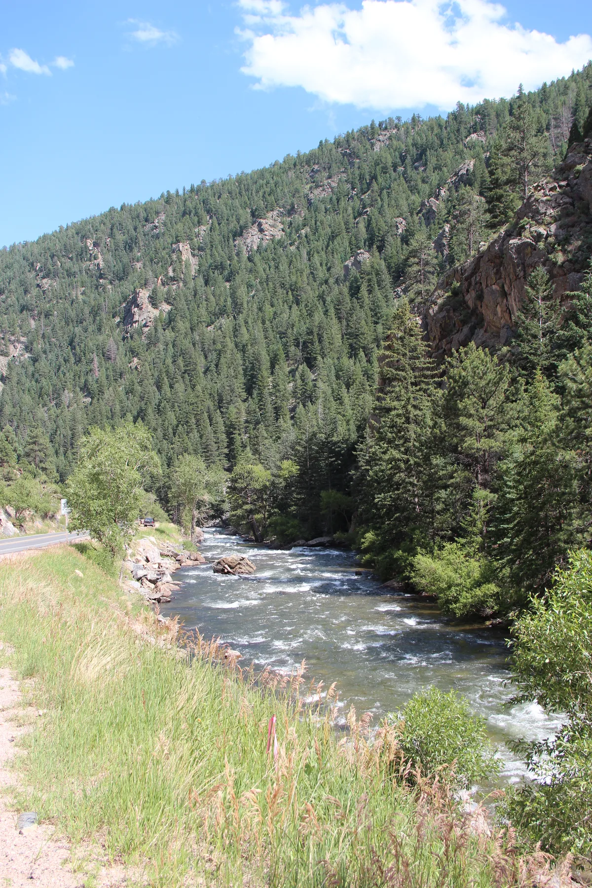 Clear Creek winds through a steep, forested canyon beside a narrow roadside pullout, with sunlit pine-covered slopes rising sharply on both sides. Bright morning light catches the rippling water and the rocky cliffs under a clear blue sky.